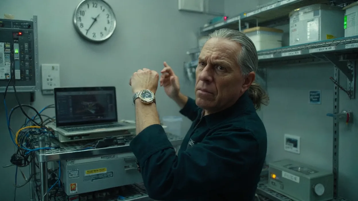 A sysadmin in a server room checks his wristwatch against the wall clock, a laptop on the equipment cart behind him.