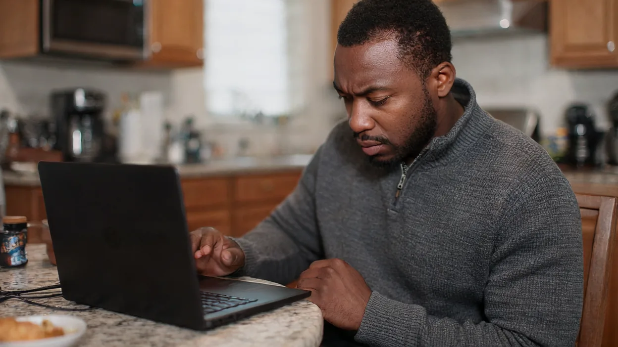 A frowning man in a gray quarter-zip sweater types on a black laptop at a granite kitchen counter.