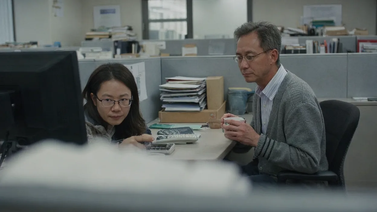 A help desk technician sits beside an older accountant at his desk in a cubicle office.